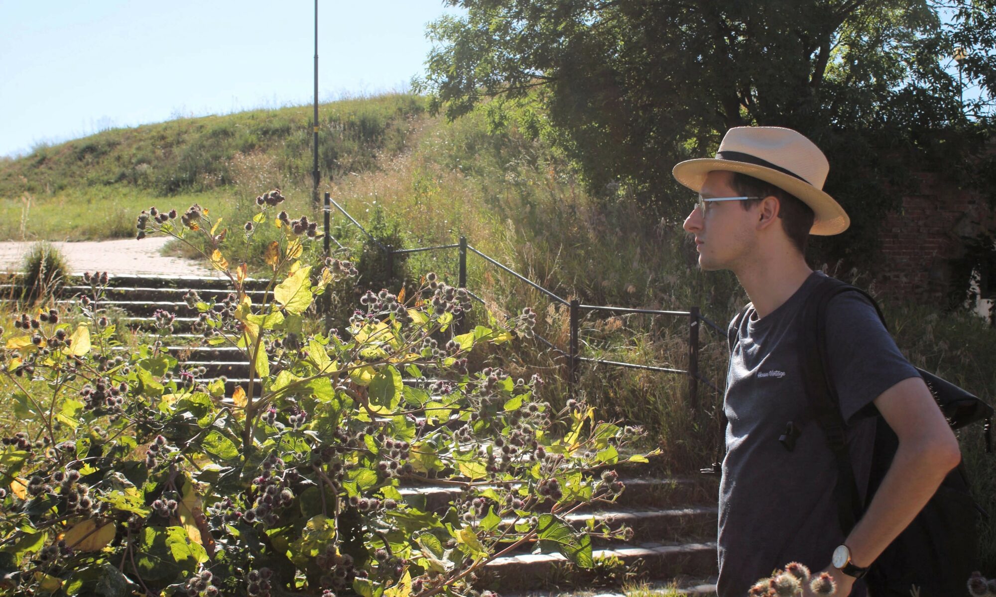 Maciej Wcisło w kapeluszu panama i podkoszulku na tle zielonych wałów ziemnych nowożytnych fortyfikacji Gdańska (okolice Kamiennej Grodzy). Badacz podczas audytu terenowego krajobrazu historycznego. Lato 2022.