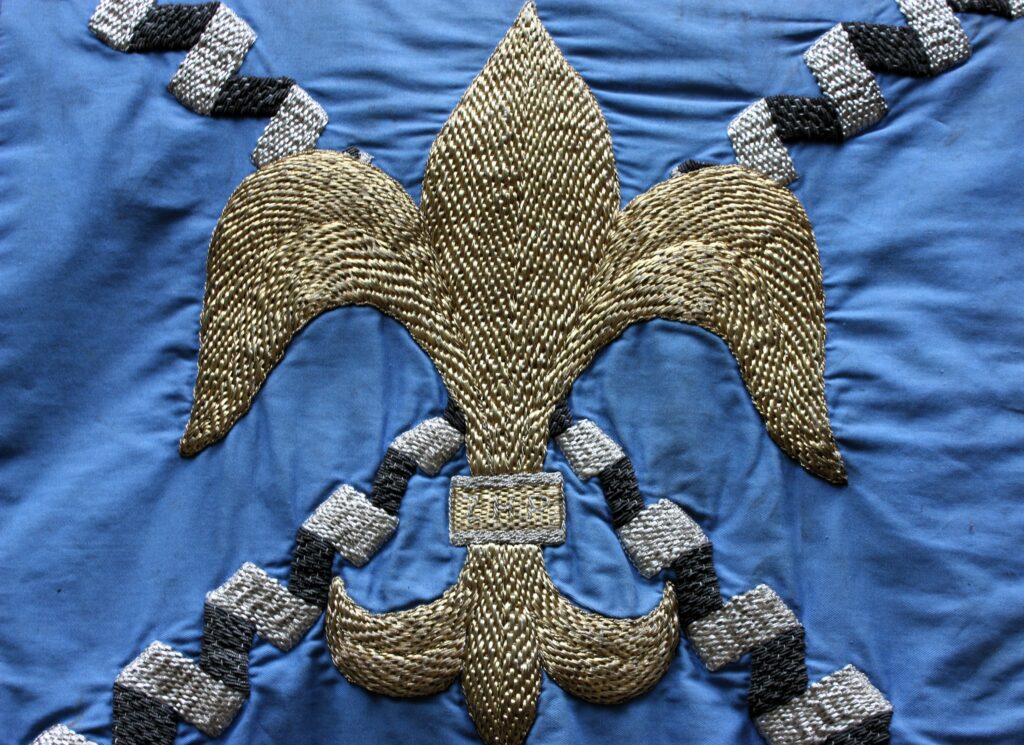 Zbliżenie na detal historycznego płomienia (proporca) do werbla. Na błękitnym tle haftowana złotą i srebrną nicią lilijka ZHP. Wyrób rzemiosła artystycznego Spółdzielni im. St. Wyspiańskiego z 1970 roku. Widoczna faktura szlachetnych nici i wykończenie bajorkiem.