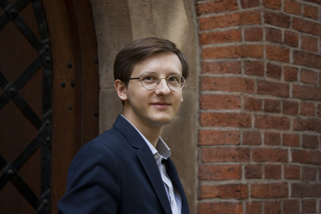 Maciej Wcisło w eleganckiej marynarce i koszuli na tle ceglanej ściany Collegium Maius w Ogrodzie Profesorskim UJ. W tle widoczne zabytkowe drewniane drzwi z okuciami i fragment kamiennego portalu. Zdjęcie wizerunkowe z kampanii wyborczej do Wydziałowej Rady Samorządu Studentów Wydziału Polonistyki UJ (maj 2019).