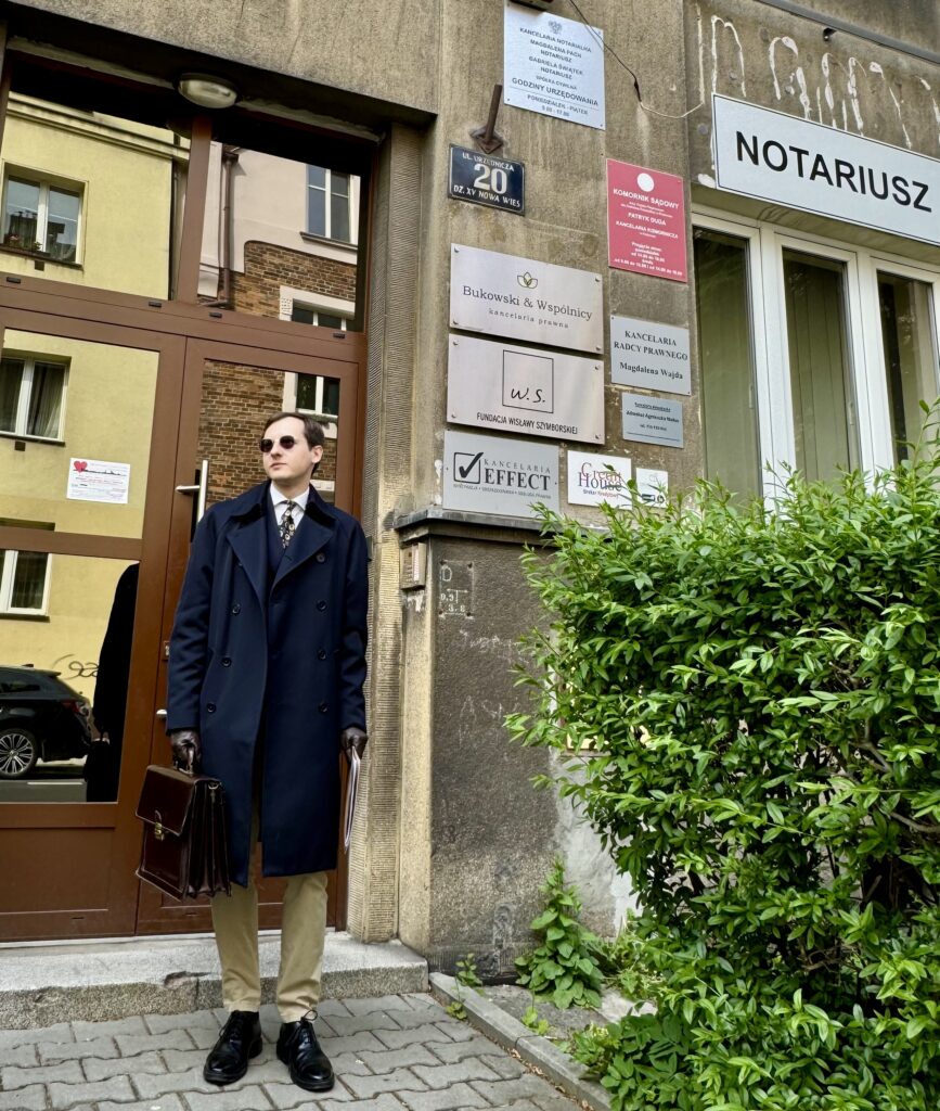Maciej Wcisło w eleganckim dwurzędowym garniturze, trzymający skórzaną aktówkę i akt notarialny, stoi przed modernistyczną kamienicą przy ulicy Urzędniczej 20 w Krakowie. W tle widoczne szyldy kancelarii notarialnej oraz biura Fundacji Wisławy Szymborskiej na krakowskiej Krowodrzy.